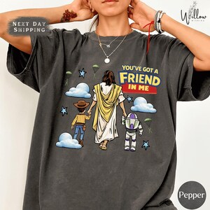 A person wearing a gray t-shirt with a graphic design depicting a religious figure, a boy, and a robot, along with the text "You've got a FRIEND in me".