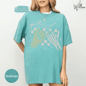 A woman wearing a turquoise t-shirt with a graphic design featuring a skeleton hand and bats, along with the text "Seafoam" and "Next Day Shipping".