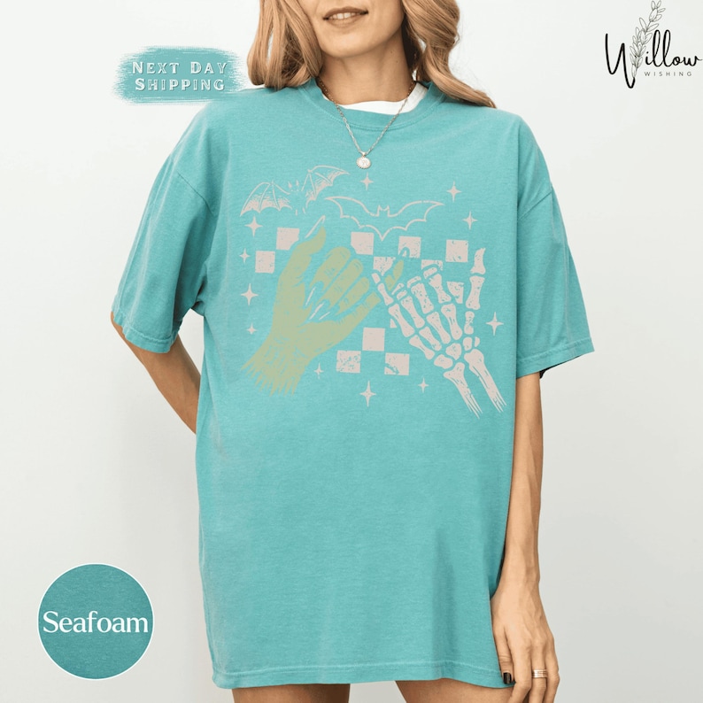 A woman wearing a turquoise t-shirt with a graphic design featuring a skeleton hand and bats, along with the text "Seafoam" and "Next Day Shipping".