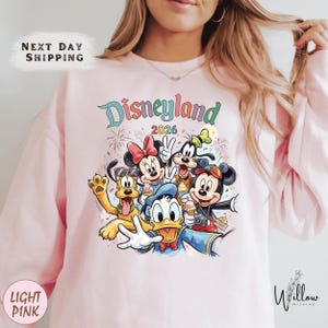 A woman wearing a pink sweatshirt with a colorful graphic of Disney characters from Disneyland.