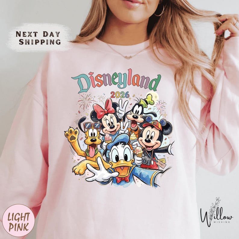 A woman wearing a pink sweatshirt with a colorful graphic of Disney characters from Disneyland.