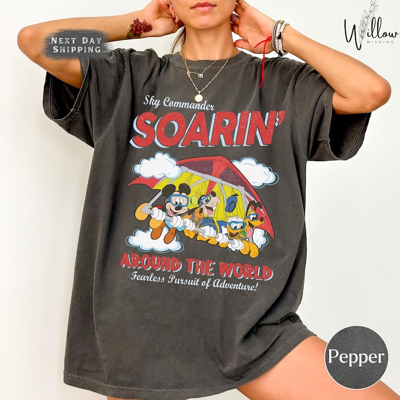 A person wearing a black t-shirt with a graphic design of Mickey Mouse and Donald Duck on a parachute, along with the text "Sky Commander SOARIN' AROUND THE WORLD Fearless Pursuit of Adventure!"