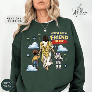 A woman wearing a green sweatshirt with a graphic design depicting a woman holding hands with a child and a robot, along with the text "You've got a FRIEND in ME".