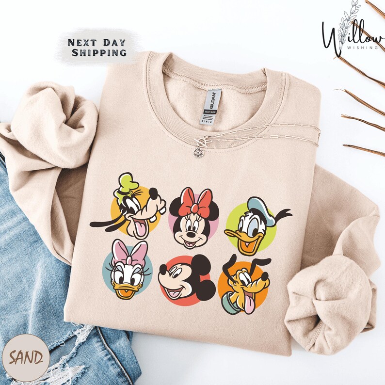 A beige sweatshirt with a graphic of Disney character faces, including Goofy, Minnie Mouse, Donald Duck, Mickey Mouse, Donald Duck, and Pluto, arranged in a row.