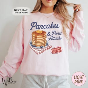 May include: Pale pink sweatshirt featuring the text "Pancakes & Panic Attacks" in navy blue. The design includes a stack of pancakes with butter and syrup. The sweatshirt also has the text "BUY ONE GET ONE".