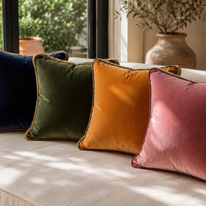 May include: Four square velvet throw pillows arranged in a row. The pillows are navy blue, olive green, mustard yellow, and dusty rose pink. Each pillow features a contrasting piped edge trim that outlines the square shape of the cushion cover.