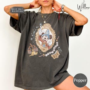 May include: Dark gray t-shirt featuring a framed illustration of Mickey Mouse in a wizard's hat and robe. The word "FANTASY" is spelled out around the frame. The shirt also has the text "NEXT DAY SHIPPING".
