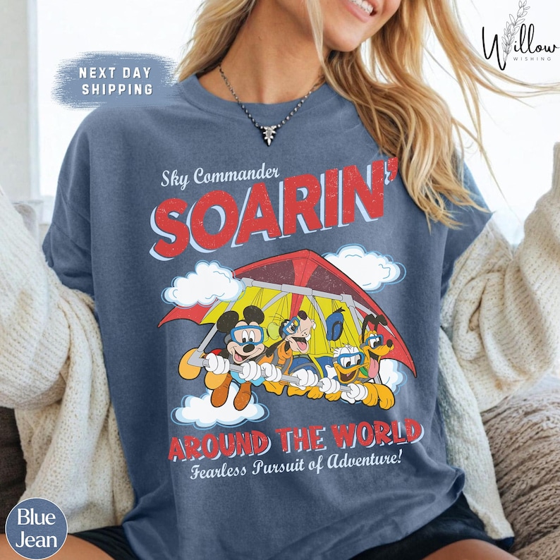 A woman wearing a blue t-shirt with a cartoon image of Mickey Mouse and Donald Duck on a hang glider, along with the text "Sky Commander SOARIN' AROUND THE WORLD Fearless Pursuit of Adventure!"