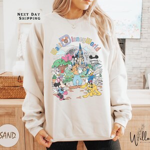 A woman wearing a white sweatshirt with a colorful graphic of Disney characters and landmarks, including Mickey Mouse and the castle of Disneyland.