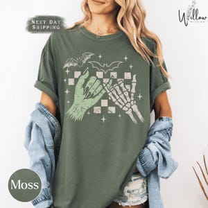 A woman wearing a green t-shirt with a graphic design of a skeleton hand and bat wings, along with the text "Moss" and "Next day shipping".