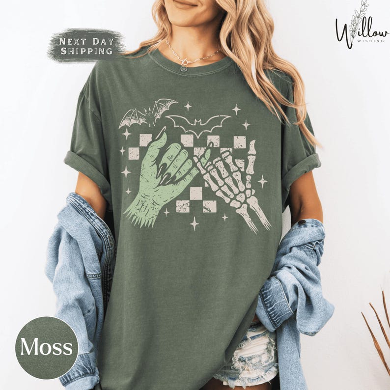 A woman wearing a green t-shirt with a graphic design of a skeleton hand and bat wings, along with the text "Moss" and "Next day shipping".