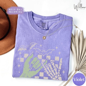 a purple t-shirt with a graphic design of a skeleton hand and bats, along with a brown hat and a fan.