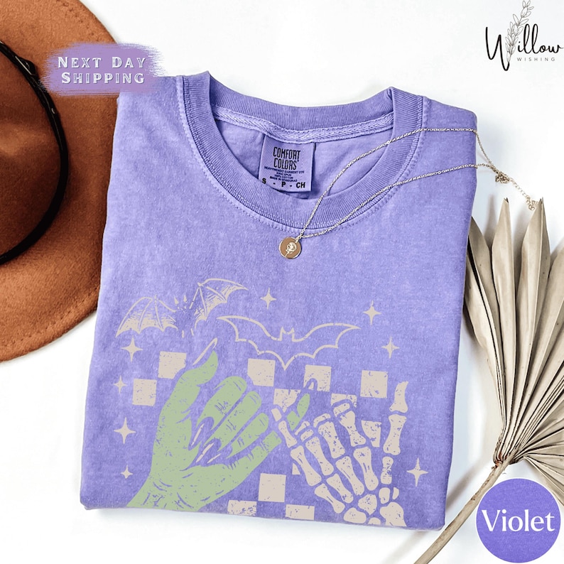 a purple t-shirt with a graphic design of a skeleton hand and bats, along with a brown hat and a fan.