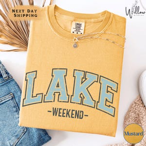 a yellow t-shirt with the word "LAKES" printed on it, along with the text "WEEKEND" below it. The t-shirt is displayed on a white background, and there are some jeans and a necklace visible in the image.