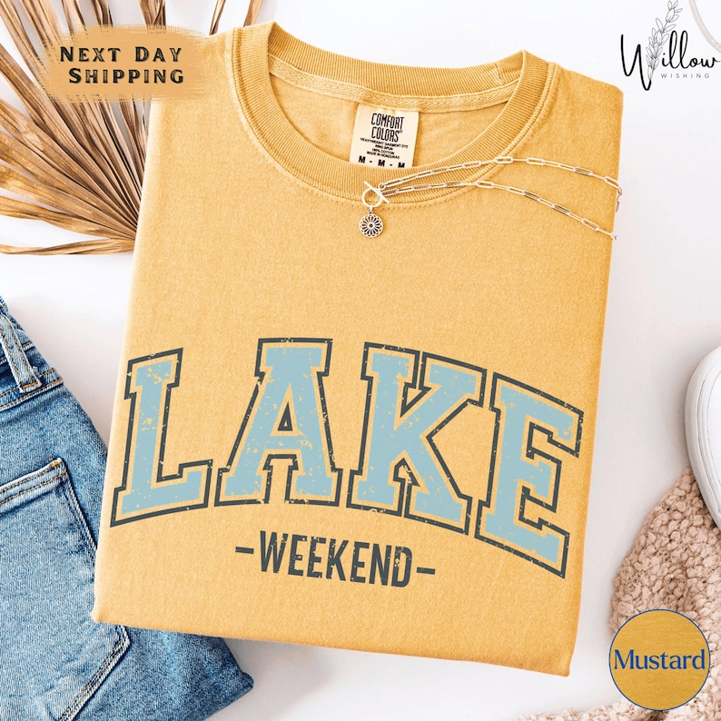 a yellow t-shirt with the word "LAKES" printed on it, along with the text "WEEKEND" below it. The t-shirt is displayed on a white background, and there are some jeans and a necklace visible in the image.