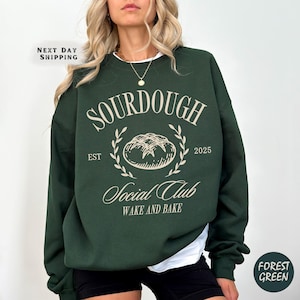 May include: A dark green sweatshirt with the text "SOURDOUGH Social Club Wake and Bake" and a graphic of a loaf of bread with the text "EST 2025"