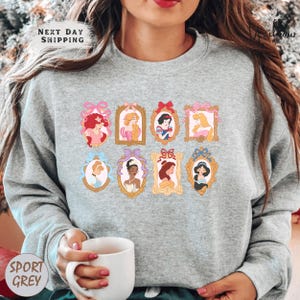 A woman wearing a gray sweatshirt with a graphic of Disney princesses in frames. She is holding a white mug in her hand.