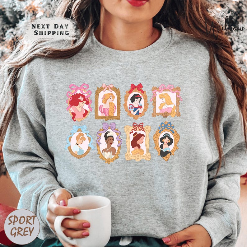 A woman wearing a gray sweatshirt with a graphic of Disney princesses in frames. She is holding a white mug in her hand.
