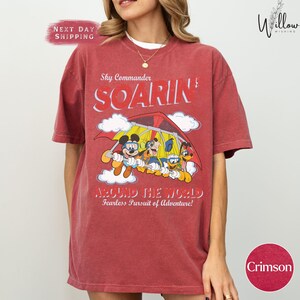 A woman wearing a red t-shirt with a cartoon image of Mickey Mouse and other characters, along with the text "Sky Commander SOARIN Around the World Fearless Pursuit of Adventure!" printed on it.