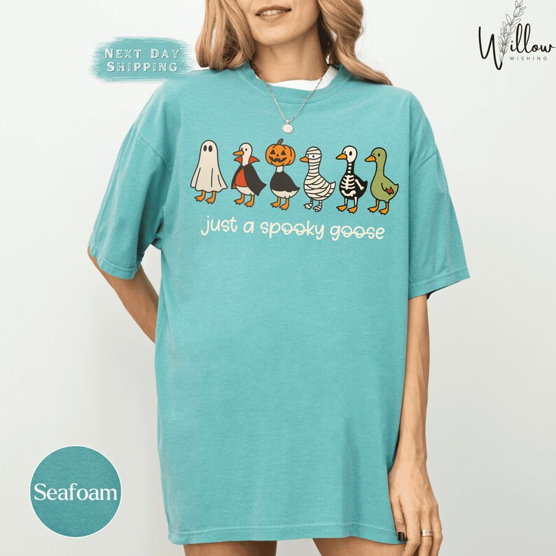 A woman wearing a turquoise t-shirt with a graphic design of various cartoon animals, including a ghost, pumpkin, mummy, and geese, along with the text "Just a spooky goose".