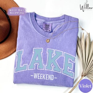 a purple t-shirt with the word "LAKES" printed on it, along with the text "WEEKEND" below it. The t-shirt is laid out on a white surface, and there is a brown hat and a fan visible in the background.