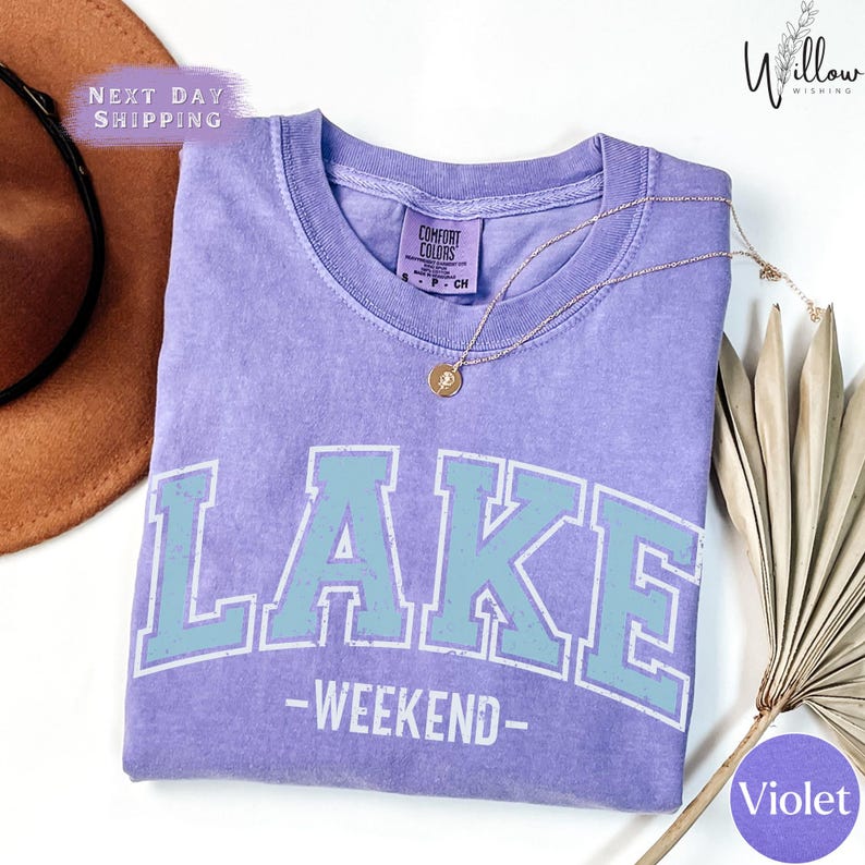 a purple t-shirt with the word "LAKES" printed on it, along with the text "WEEKEND" below it. The t-shirt is laid out on a white surface, and there is a brown hat and a fan visible in the background.