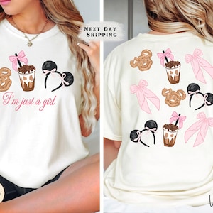 May include: Cream-coloured t-shirt with a graphic design featuring a pretzel, a drink with a straw, and a Minnie Mouse ear headband. The text "I'm just a girl" is written in pink script. The back of the shirt has the same design.