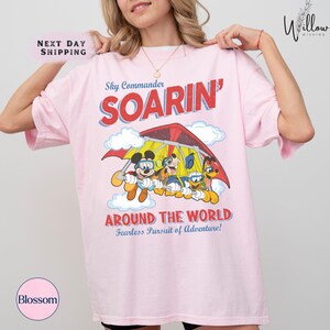 A woman wearing a pink t-shirt with a cartoon image of Mickey Mouse and other characters, along with the text "Sky Commander SOARIN' AROUND THE WORLD Fearless Pursuit of Adventure!"