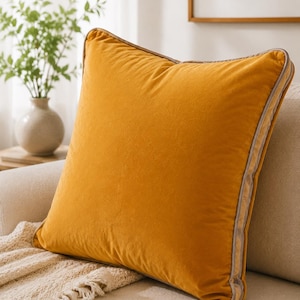 May include: A square mustard yellow velvet throw pillow featuring a smooth, solid-colored front and back. The pillow is finished with a light beige piping trim along all four edges, providing a clean, structured border to the soft fabric surface.