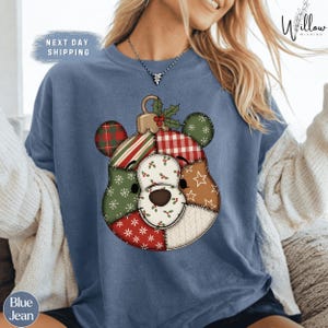 Peut inclure: Sweat-shirt bleu marine avec un motif d'ornement d'ours en patchwork. L'ours présente divers motifs, notamment des carreaux, des rayures et des flocons de neige. Le texte "NEXT DAY SHIPPING" est visible en haut à gauche.