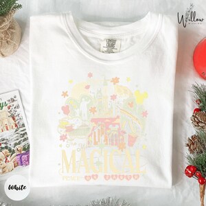 A white t-shirt with a colorful graphic design featuring various Disney-themed elements, such as Mickey Mouse, Minnie Mouse, and a castle, along with the text "The most magical planet Earth".