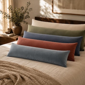 May include: A row of four long, rectangular bolster pillows arranged on a bed. The pillows are coloured in shades of light blue, terracotta, dark blue, and olive green, with a large cream-coloured pillow positioned at the back of the group.