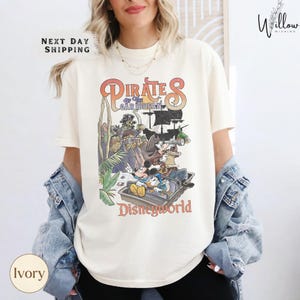 A woman wearing a white t-shirt with a graphic of Disney characters and text, standing in front of a white wall.