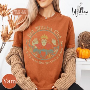 A woman wearing a t-shirt with a graphic design featuring characters from the Disney film "Bad Witches". She is also wearing a sweater and has her hair styled in loose waves.