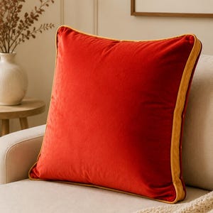 May include: A square throw pillow featuring a vibrant red velvet fabric on the front and back, accented with a contrasting mustard yellow piping trim along all four edges. The pillow has a soft, plush texture and a clean, simple square shape.