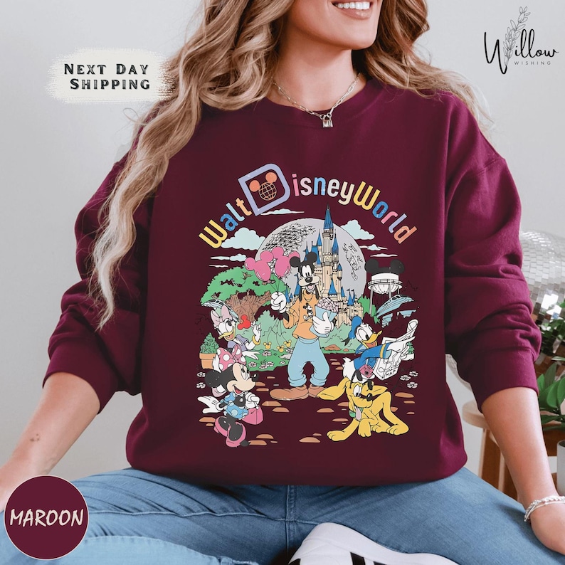 A woman wearing a burgundy sweatshirt with a colorful graphic of Disney characters and landmarks, including Mickey Mouse, Minnie Mouse, Donald Duck, and the Eiffel Tower.