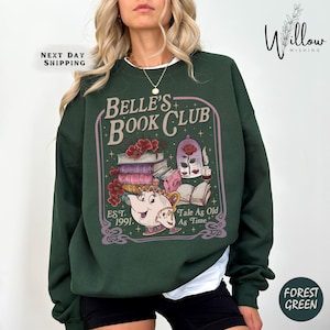 Peut inclure: Sweat-shirt vert forêt avec un motif graphique. Le motif comprend le texte "Belle's Book Club", des illustrations de livres, une rose et une théière. Le sweat-shirt porte également le texte "Tale As Old As Time" et "Est. 1991."