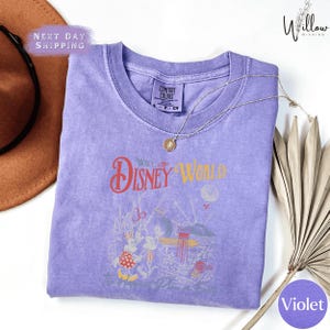 a purple t-shirt with a Disney World-themed design, including a Mickey Mouse and Minnie Mouse illustration, along with the text "Disney World" and "The Happy Place". The t-shirt is laid out on a white surface, with a brown hat and a fan visible in the background.