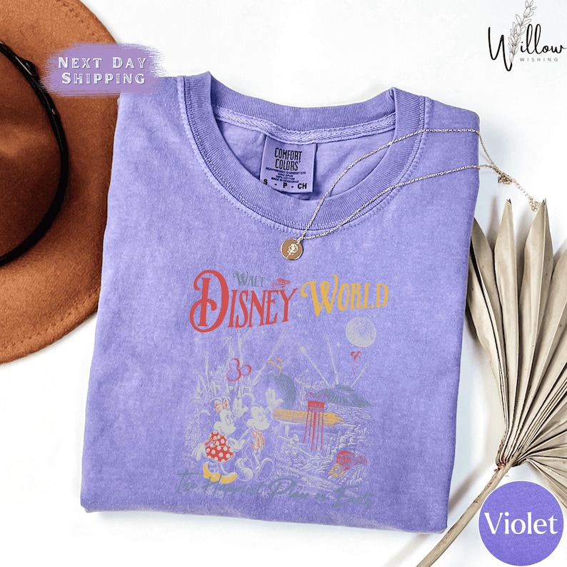 a purple t-shirt with a Disney World-themed design, including a Mickey Mouse and Minnie Mouse illustration, along with the text "Disney World" and "The Happy Place". The t-shirt is laid out on a white surface, with a brown hat and a fan visible in the background.