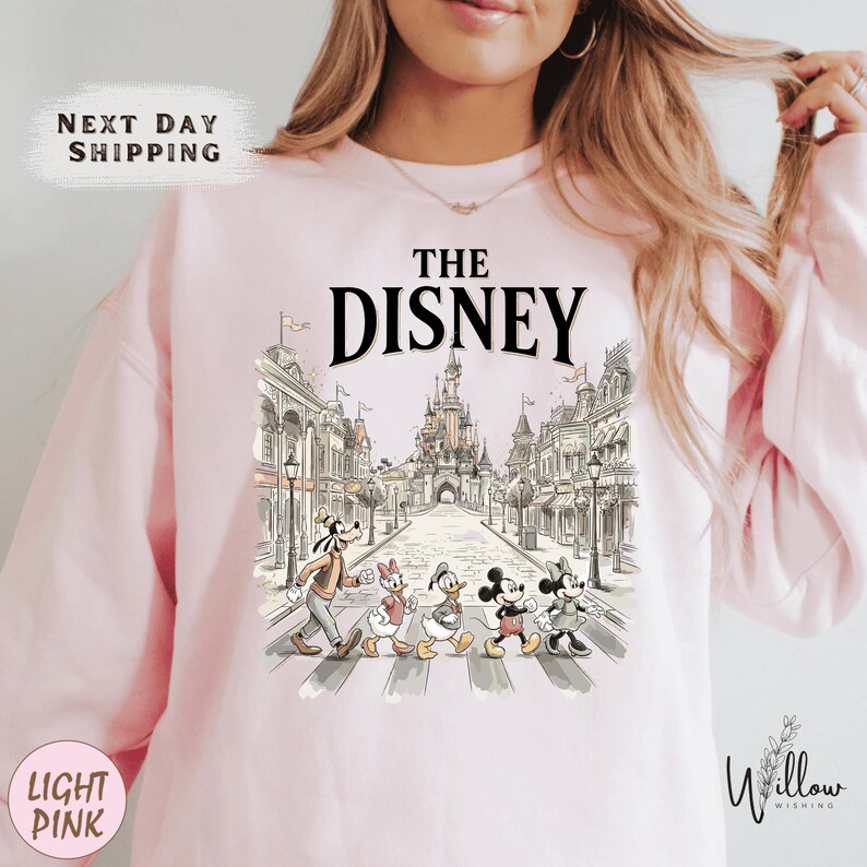 A woman wearing a pink sweatshirt with a graphic of Disney characters crossing the street.