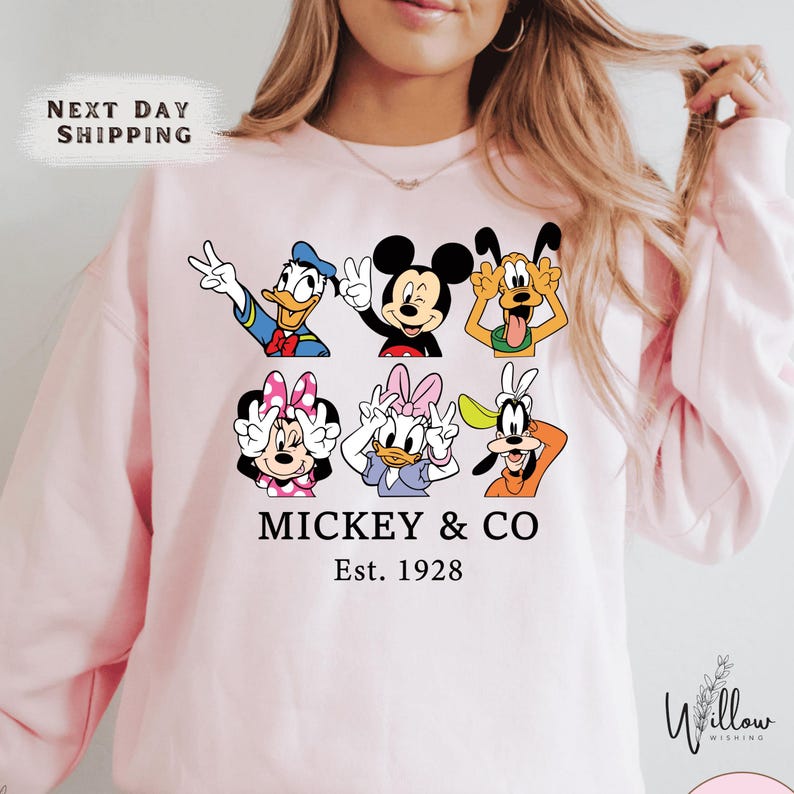 A woman wearing a pink sweatshirt with a graphic design featuring various Disney characters, including Mickey Mouse and Donald Duck.