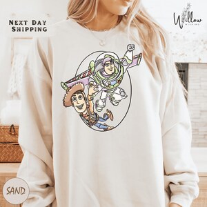 A person wearing a white sweatshirt with a cartoon illustration of two characters from the Toy Story franchise, Buzz Lightyear and Woody, depicted in a playful and energetic pose.