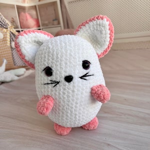 May include: A white crocheted mouse plush toy with pink accents on the ears, paws, and nose. The toy has black eyes and a stitched-on black nose and whiskers.
