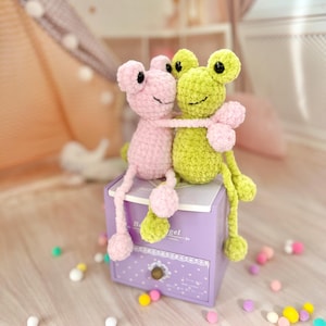 May include: Two crocheted frog plushies, one pink and one green, are hugging each other. They are sitting on a purple drawer with white lace trim and the text "Baby Angel" on the front. The drawer is surrounded by colorful pom-poms.
