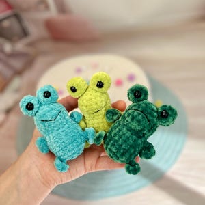 Puede incluir: Tres peluches de rana de crochet en diferentes colores: verde azulado, verde y verde lima. Las ranas tienen grandes ojos negros y están sentadas en una mano.