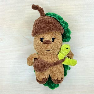 Könnte beinhalten: Handgefertigte Plüsch-Eichel-Figur mit brauner Kappe, beigem Körper und grünen Blättern. Das Plüschtier hat ein lächelndes Gesicht und hält einen grünen Blattzweig. Das Spielzeug ist aus weichem, strukturiertem Garn gefertigt.