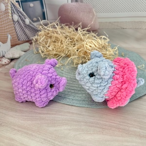 May include: Two handmade crochet pig toys. One is solid purple, the other is gray and pink. The pigs are on a gray mat with straw in the background. The toys are small and have black button eyes.
