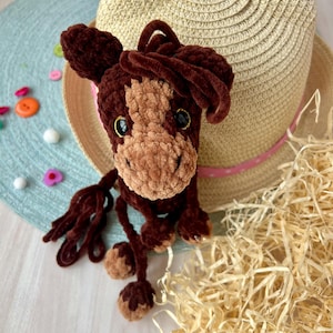 Może przedstawiać: Ręcznie robiona brązowo-beżowa zabawka w kształcie konia z czarnymi oczami i puszystą grzywą. Koń jest umieszczony przed słomkowym kapeluszem z różową wstążką. Ozdobne guziki i słoma są rozrzucone wokół zabawki.