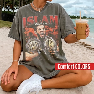 May include: Dark gray t-shirt featuring a graphic of Islam Makhachev with UFC championship belts. The shirt has the word "ISLAM" in red letters and the text "Comfort COLORS" in a red rectangle.