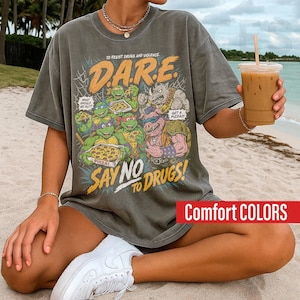 May include: A charcoal grey short-sleeve t-shirt featuring a graphic print of four cartoon turtles and two anthropomorphic characters eating pizza. The text reads D.A.R.E. TO RESIST DRUGS AND VIOLENCE, SAY NO TO DRUGS, WHAT SHOULD WE DO?, and GET A PIZZA.